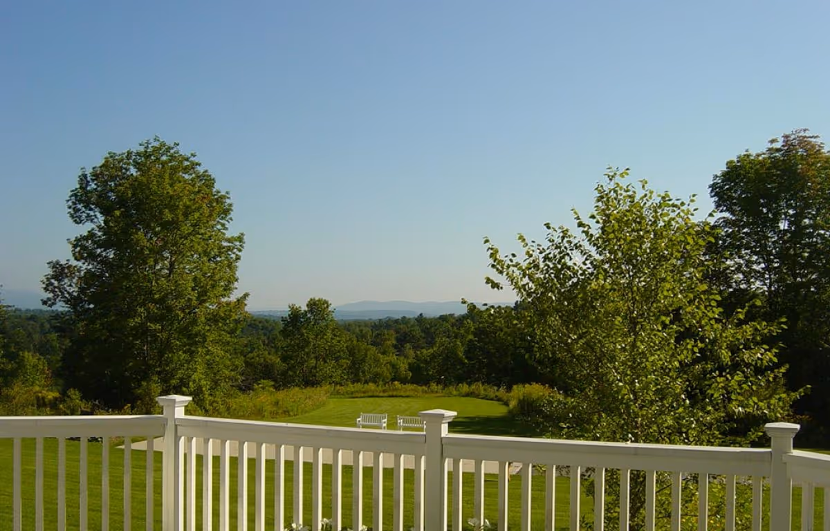 View from a white wooden balcony railing overlooking a green lawn with two white benches, surrounded by trees and distant hills under a clear blue sky.