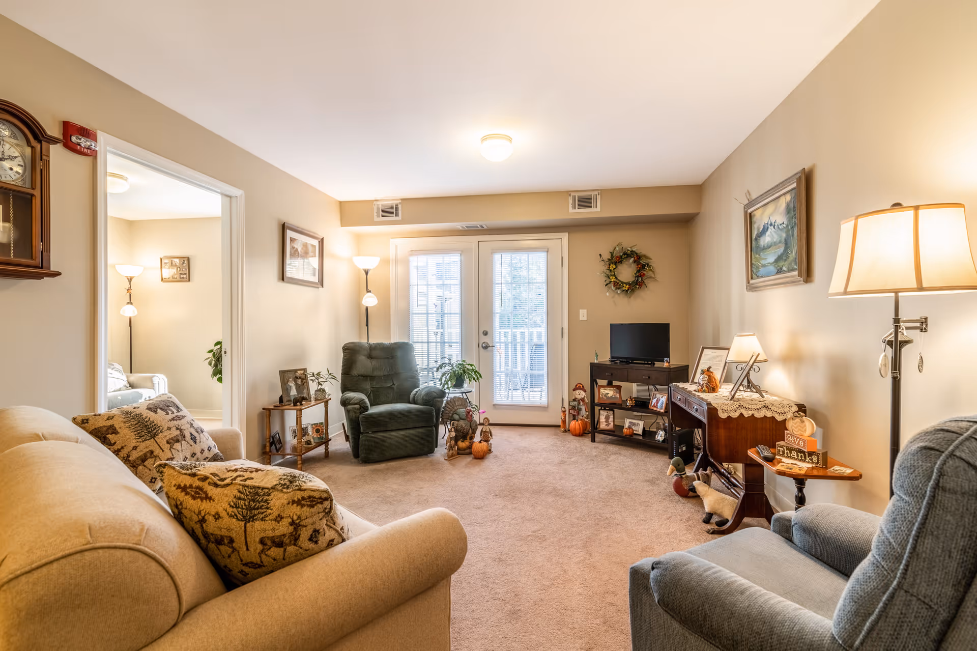 A cozy living room with beige walls and carpet, featuring a beige sofa with patterned cushions, a green recliner chair, and a blue armchair. There is a wooden grandfather clock on the left wall, a small side table with plants and picture frames, a TV on a stand, and a wooden console table with lamps and decorative items. French doors with blinds lead to an outdoor balcony. The room is warmly lit with floor and table lamps, and decorated with framed artwork and a wreath.
