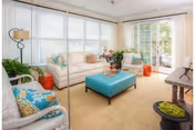 A bright and airy living room with large windows covered by white blinds, a white sofa with colorful cushions, a turquoise ottoman in the center, two armchairs with patterned cushions, and various plants and decorative items. There is a glass door leading to an outdoor area.