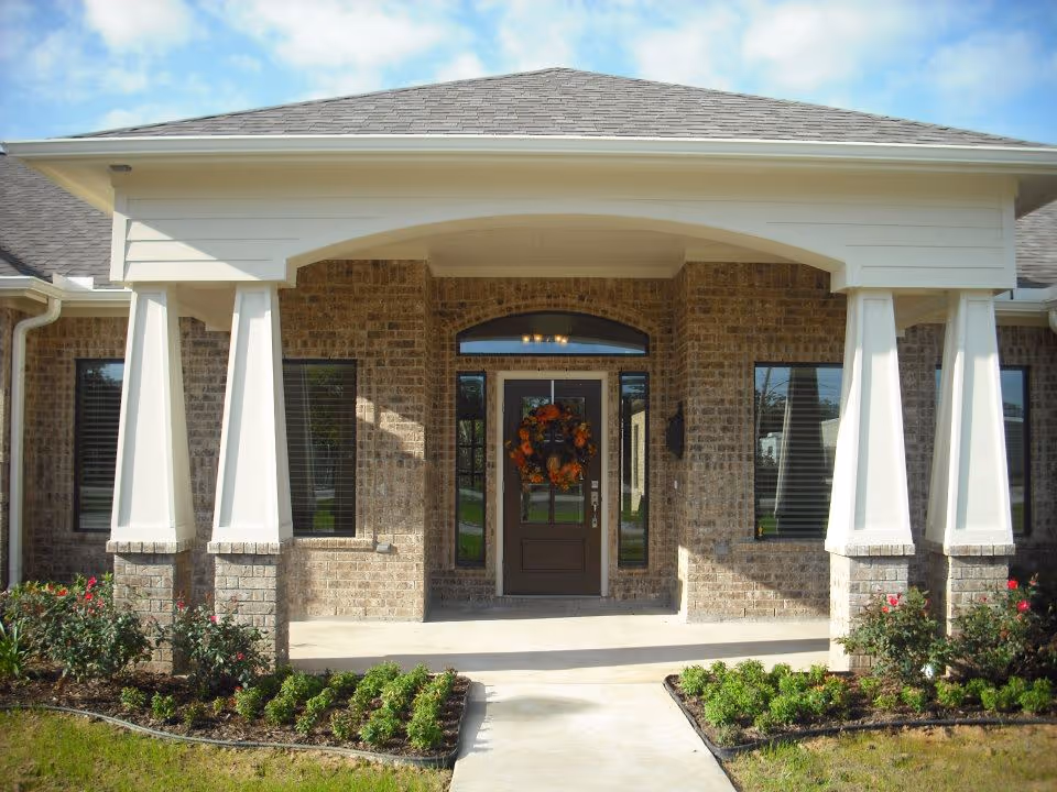 Front entrance of a brick building with a covered porch supported by white columns. The door has a decorative wreath with orange and red flowers. There are windows on either side of the door and small landscaped garden beds with green shrubs and flowers in front.
