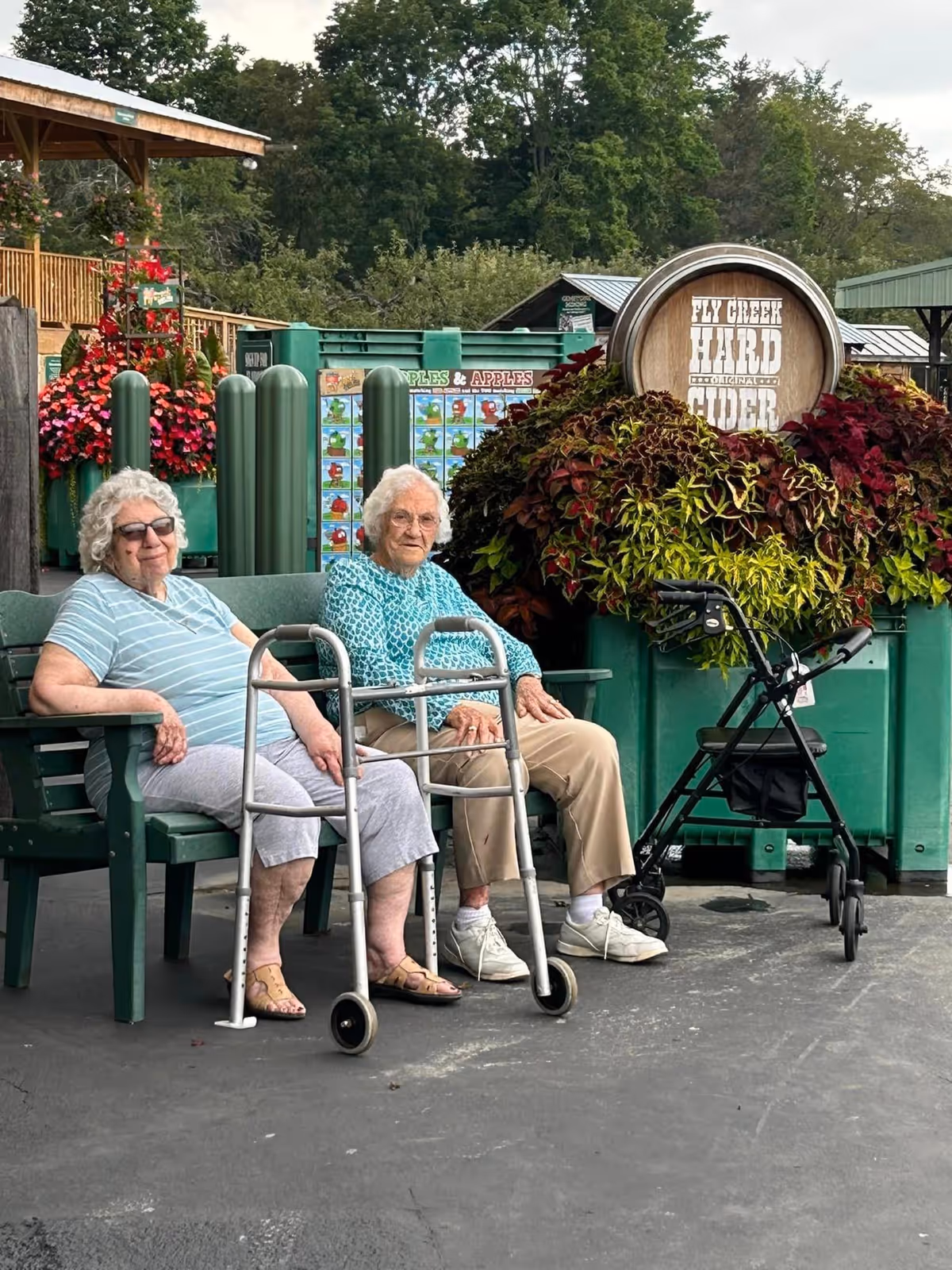 Two elderly women sitting on a green bench outdoors. One woman is wearing a striped shirt and sunglasses, and the other is wearing a patterned blue top. Both have walkers in front of them. Behind them is a large planter with colorful foliage and a wooden barrel sign that reads 'Fly Creek Hard Cider'. There are trees and a wooden structure in the background.