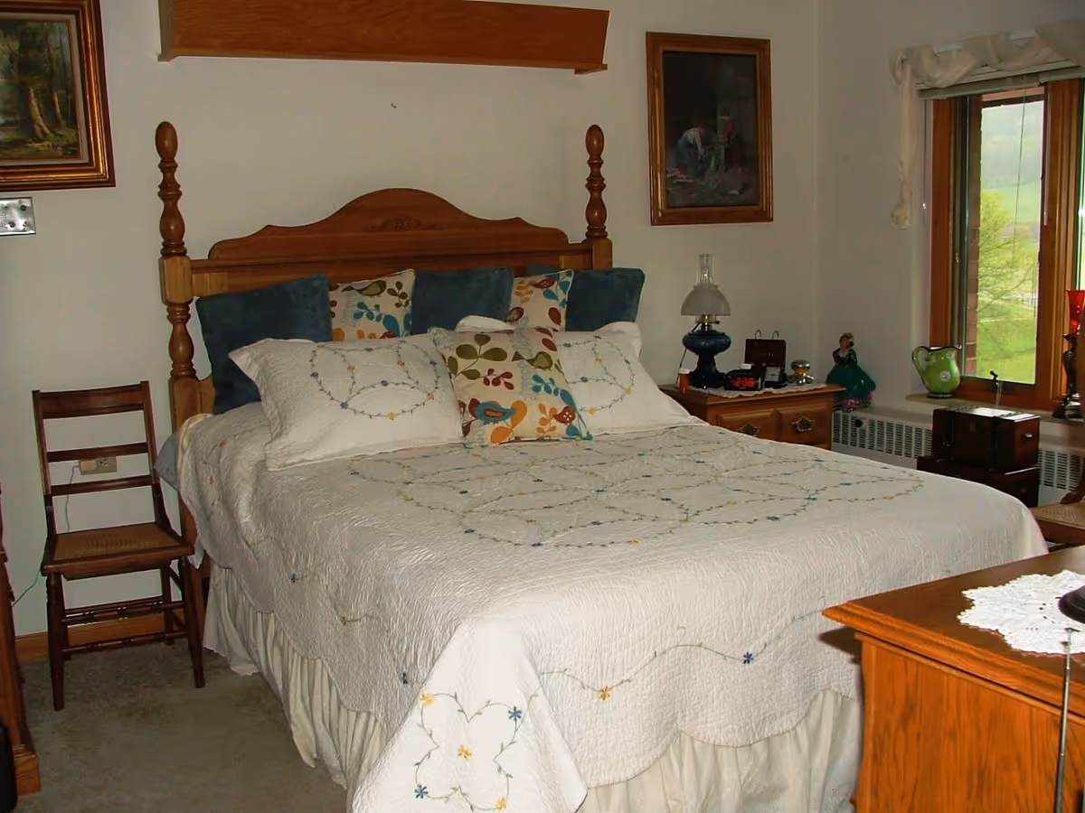 A cozy bedroom with a wooden bed frame and a white quilted bedspread decorated with floral patterns. The bed has multiple pillows with floral and solid blue covers. There is a wooden chair to the left of the bed and a wooden nightstand to the right with a vintage lamp, clock, and decorative items. A window with a view of greenery is on the right wall, and two framed paintings hang on the walls.