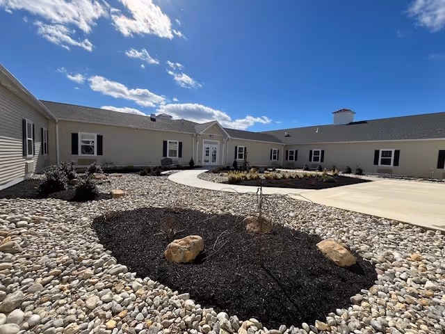 Exterior view of a single-story building with beige siding and black shutters under a partly cloudy blue sky. The foreground features a landscaped area with rocks, mulch, and a few small plants, along with a curved concrete walkway leading to a white door entrance.