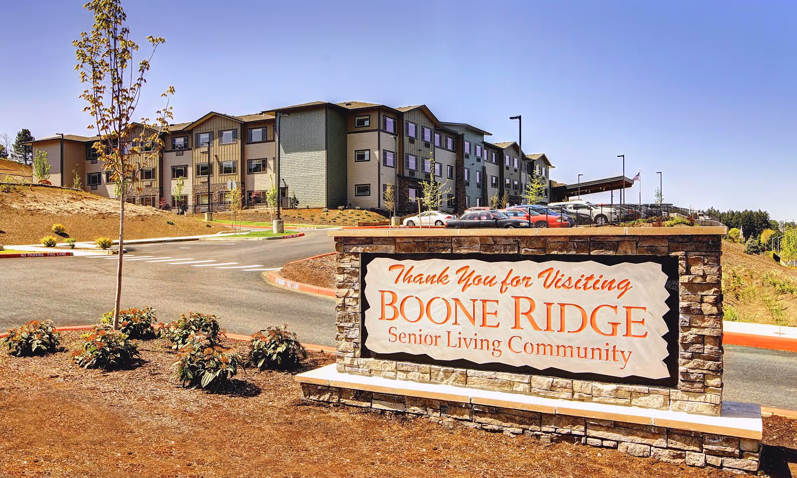 Exterior view of Boone Ridge Senior Living Community building with a stone sign in the foreground that reads 'Thank You for Visiting Boone Ridge Senior Living Community'. The building is three stories tall with multiple windows and a parking area with cars. The sky is clear and blue.
