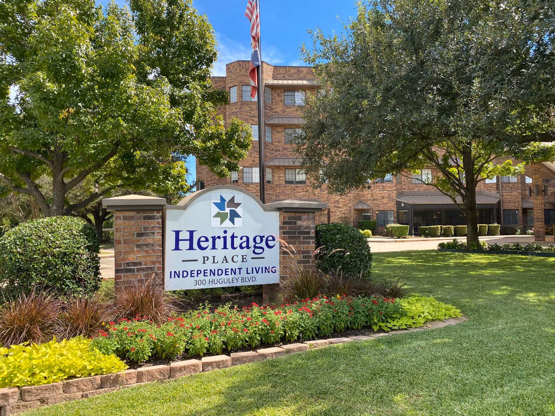 Outdoor view of Heritage Place Independent Living facility showing a brick building in the background, a landscaped garden with green grass, colorful flowers, and trees surrounding a sign that reads 'Heritage Place Independent Living 300 Huguley Blvd.' An American flag is visible behind the sign.
