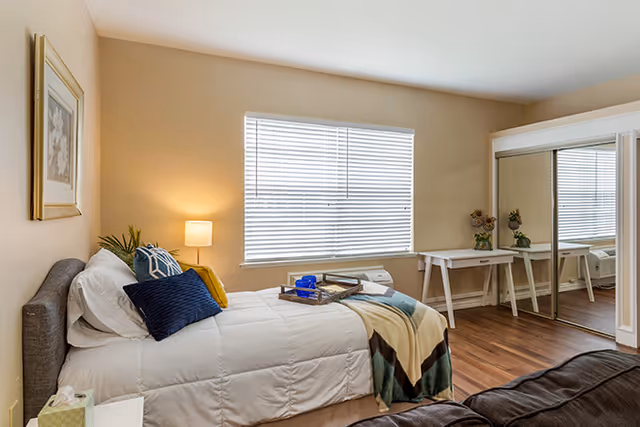 A cozy bedroom with a neatly made bed featuring white bedding, multiple pillows in blue and yellow, and a patterned throw blanket. There is a bedside table with a lamp and a plant, a large window with blinds, a white desk with decorative items, and a mirrored sliding closet door. The room has wooden flooring and beige walls.