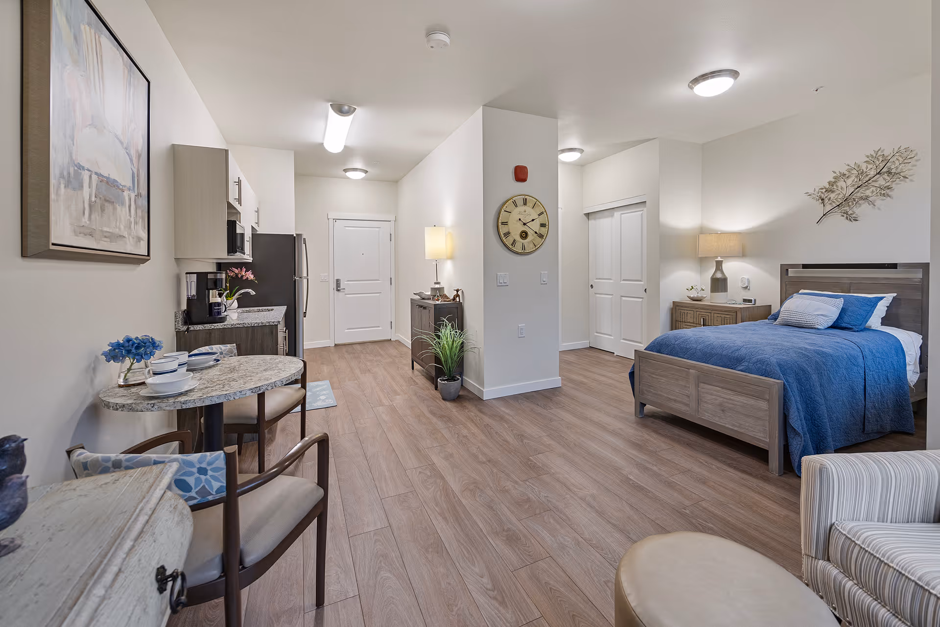 A spacious and well-lit studio apartment featuring a bed with blue bedding, a wooden nightstand with a lamp, a small dining table set with dishes and chairs, a kitchenette with appliances, and light wood flooring. The walls are decorated with a clock, a framed painting, and a metal leaf wall art. The room has a clean and modern design with neutral tones.