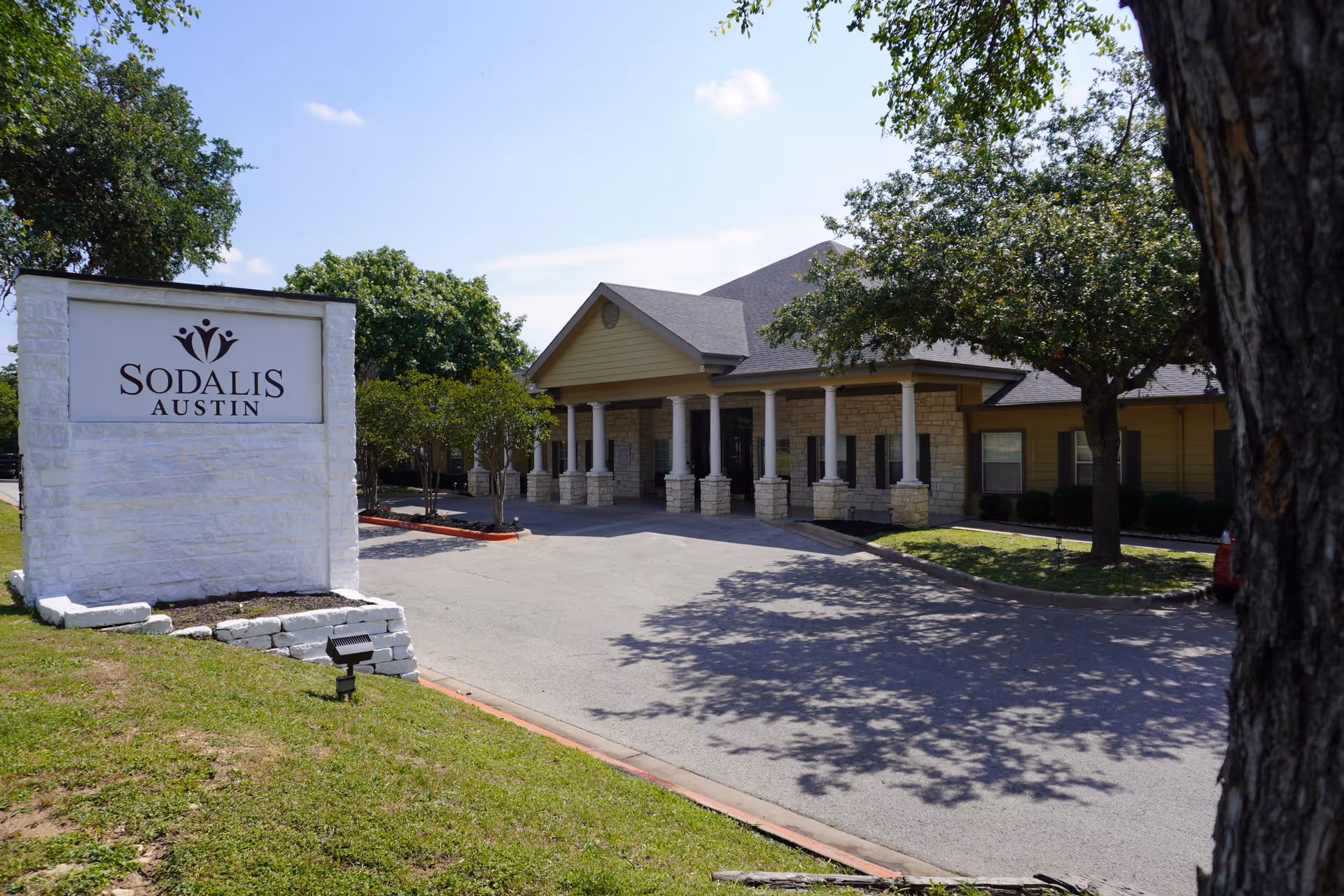 Exterior view of Sodalis Austin facility showing a single-story building with stone and beige siding, a covered entrance supported by white columns, surrounded by trees and greenery under a clear blue sky.