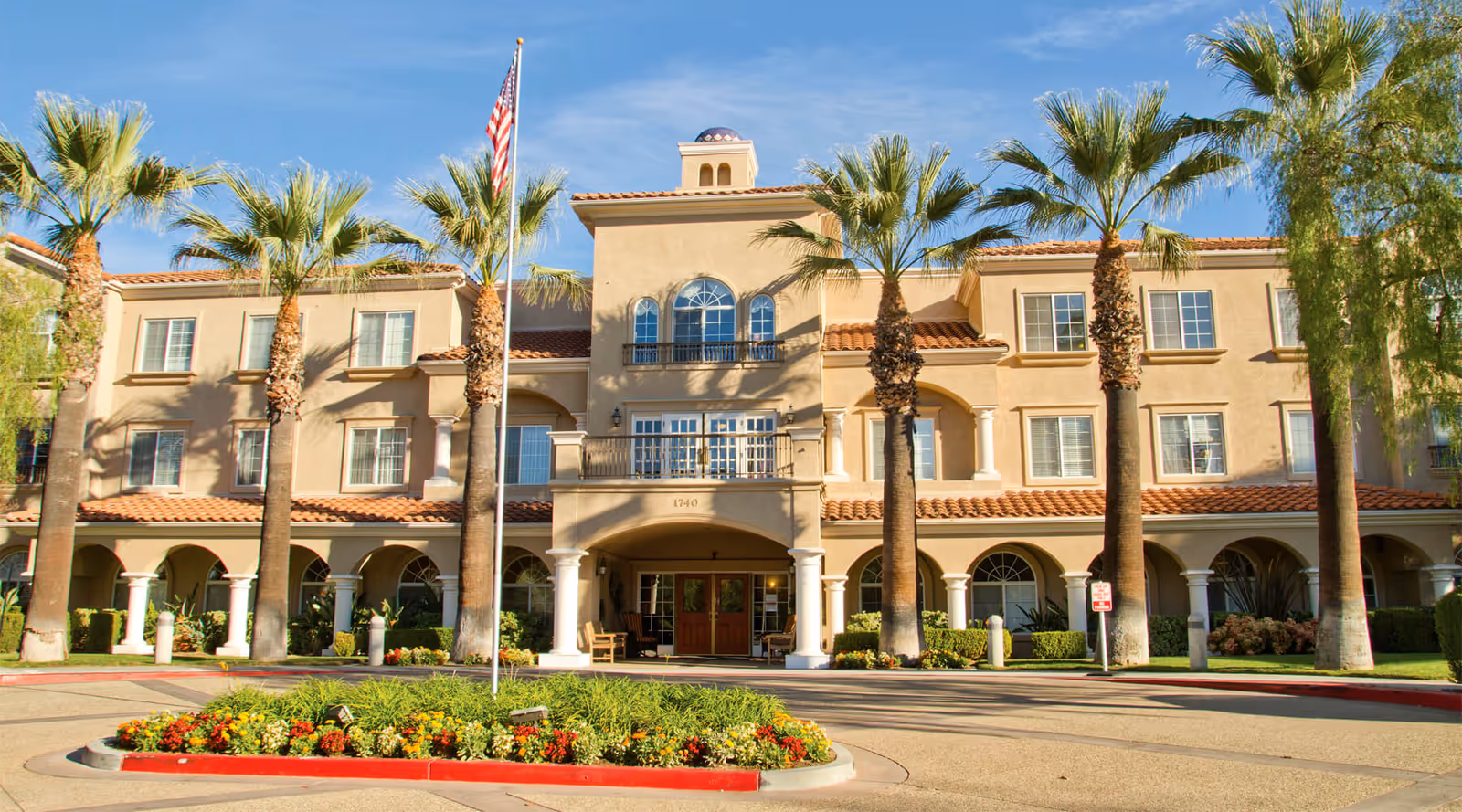 Front exterior view of a three-story senior living facility building with beige stucco walls, red tile roof, and arched windows. There are tall palm trees and a flagpole with an American flag in front of the entrance. The entrance has double wooden doors under an archway with white columns. A landscaped roundabout with flowers and greenery is in the foreground.