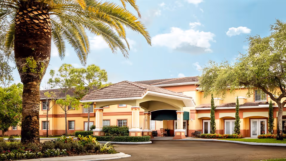 Exterior view of Wickshire Tamarac senior living facility showing a two-story building with a covered entrance, surrounded by palm trees and landscaped greenery under a partly cloudy sky.