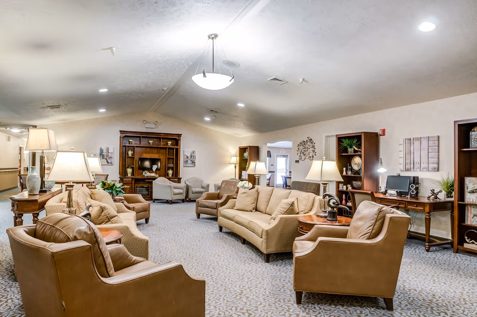 A spacious and well-lit living room area in a senior living facility with multiple beige and brown upholstered chairs and sofas arranged around wooden side tables with lamps. The room features a carpeted floor, a wooden entertainment center with a TV, bookshelves, and decorative wall art. There is also a desk with a computer and additional lighting on the right side of the room.