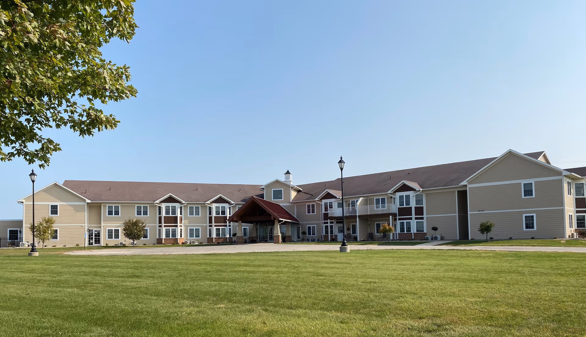 Exterior view of a two-story senior living facility building with beige siding and a brown roof, surrounded by a well-maintained green lawn and a clear blue sky. There are a few small trees and lamp posts near the building.