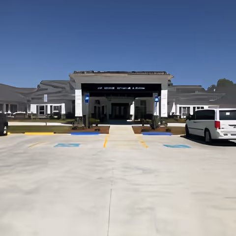 Front exterior view of a single-story senior living facility building with a covered entrance, wheelchair accessible parking spaces, and a clear blue sky.