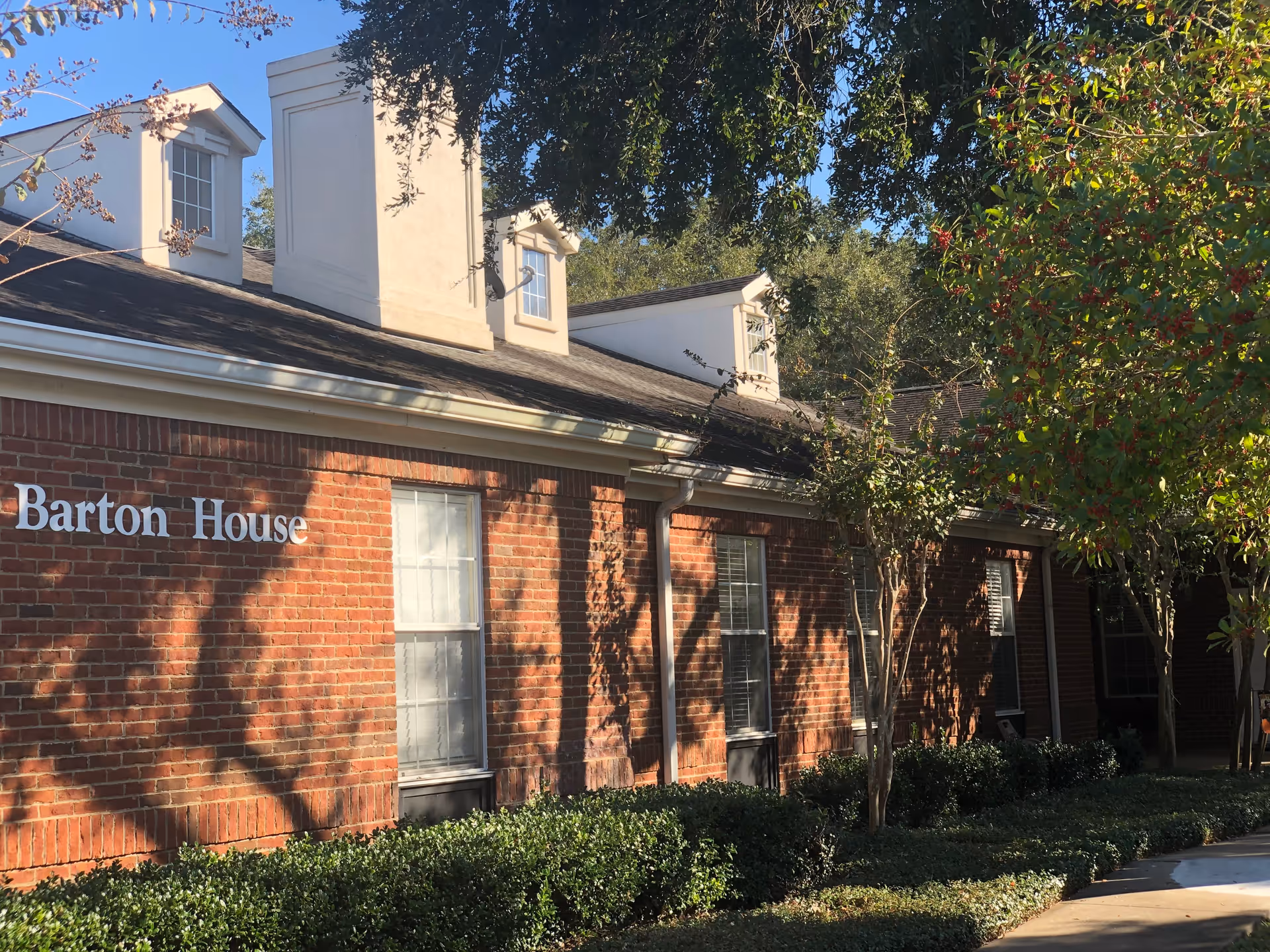 Exterior view of a brick building with white framed windows and a sign that reads 'Barton House'. The building is surrounded by green bushes and trees with sunlight casting shadows on the wall.