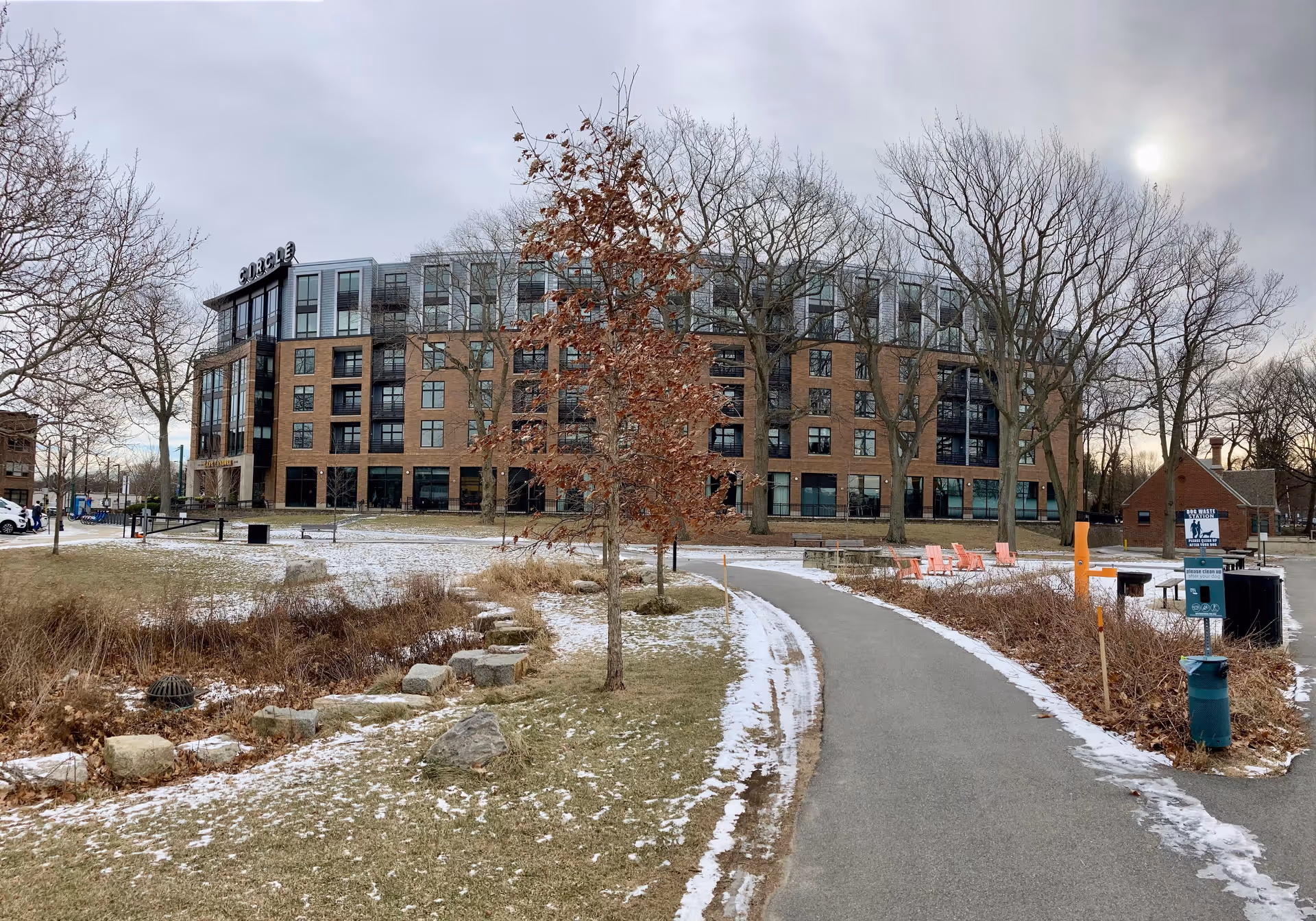 Exterior view of a multi-story senior living facility named Waterstone at the Circle, surrounded by leafless trees and a partially snow-covered lawn with a paved walking path leading towards the building. There are orange chairs and a small brick structure with signs near the path.