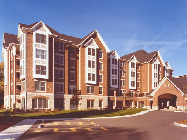 Exterior view of a multi-story senior living facility building with brick and stone facade, large windows, and a covered entrance. The building is surrounded by a paved driveway and landscaped grass areas under a clear blue sky.