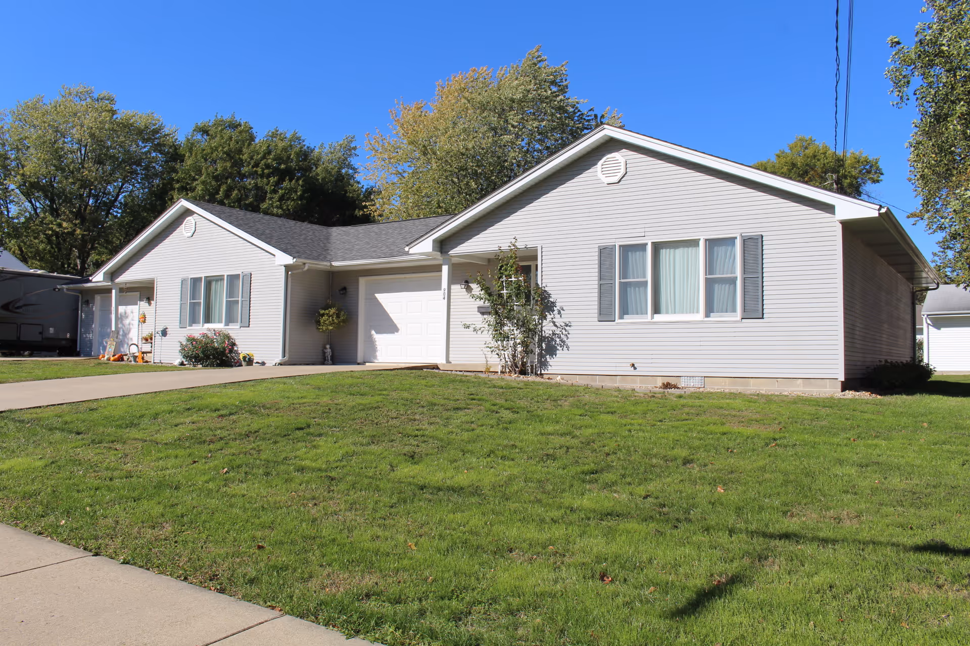 Single-story light gray building with white trim and a garage, surrounded by green grass and trees under a clear blue sky.