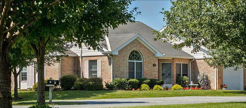 A single-story brick residential building with a gray roof, surrounded by green grass, bushes, and trees under a clear blue sky.