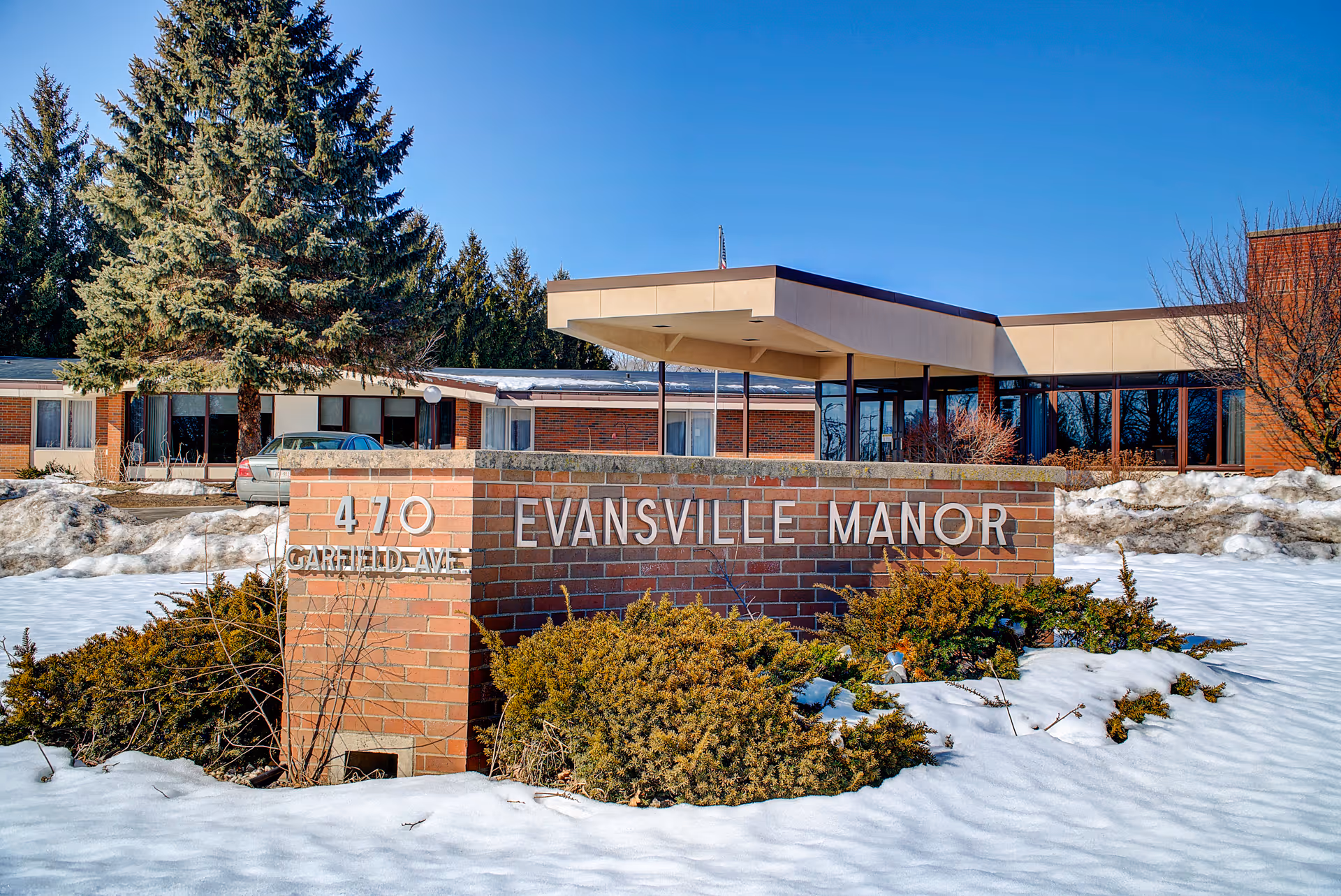 Exterior view of Evansville Manor Skilled Nursing facility with a brick sign displaying the address 470 Garfield Ave. Snow covers the ground and bushes in front of the sign, and the building is visible in the background under a clear blue sky.