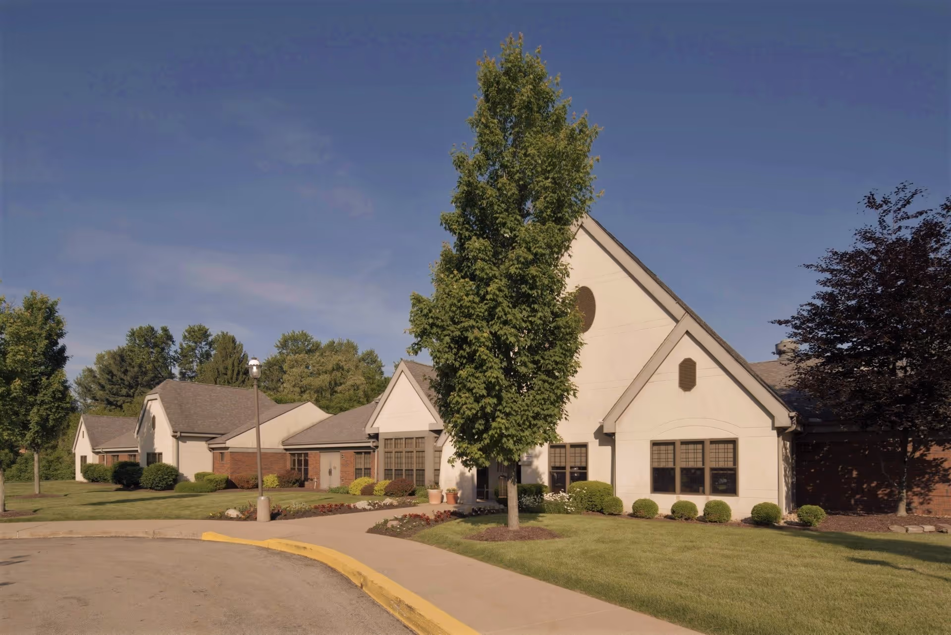 Exterior view of a single-story building with beige walls and a sloped roof, surrounded by green grass, trees, and shrubs under a clear blue sky.