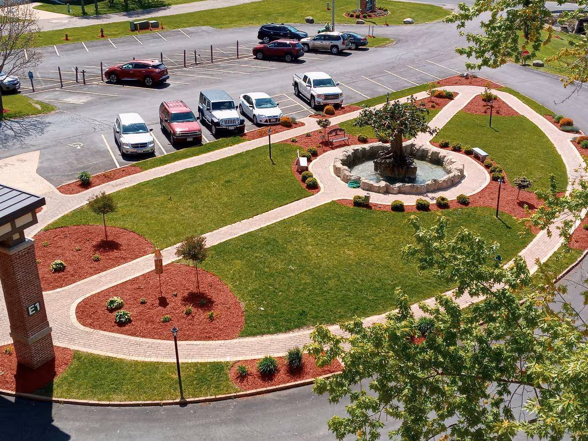 Aerial view of a landscaped garden with a central fountain, surrounded by walking paths and parking spaces, featuring green grass and decorative plants.