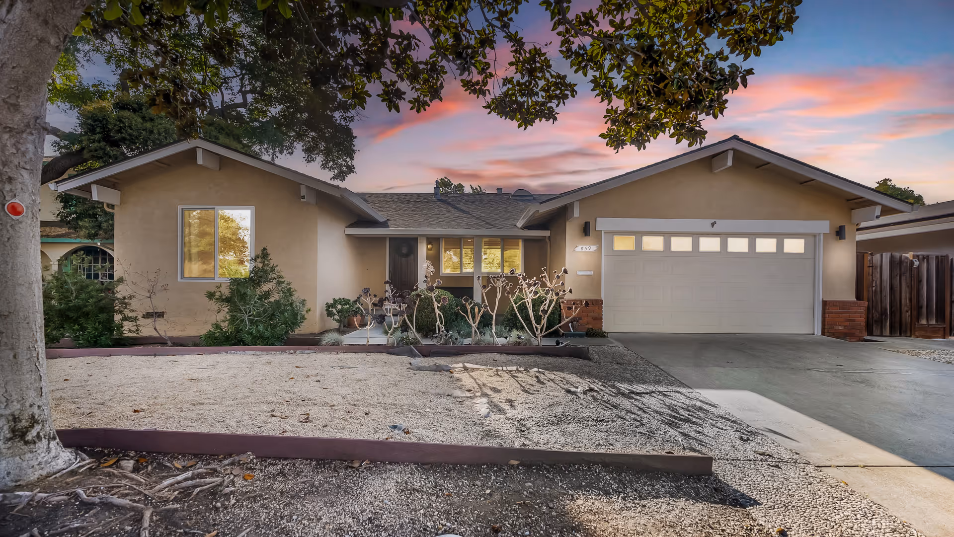 Front exterior view of a single-story beige house with a two-car garage, a driveway, and a landscaped front yard with gravel and some plants. The sky is colorful with a sunset or sunrise.