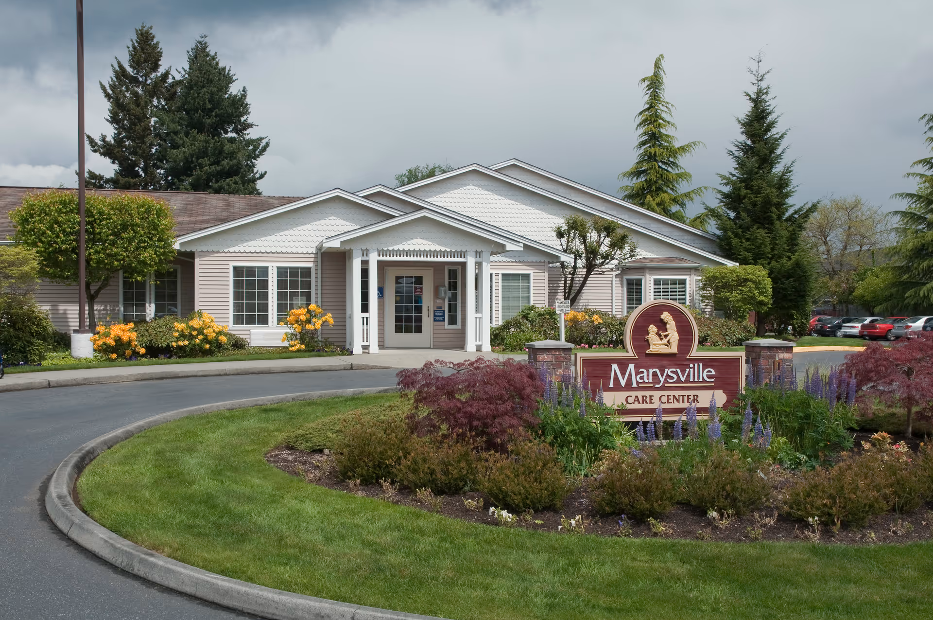 Exterior view of Marysville Care Center building with a landscaped garden and a sign displaying the facility's name in front. The building is single-story with white trim and surrounded by trees and shrubs under a cloudy sky.