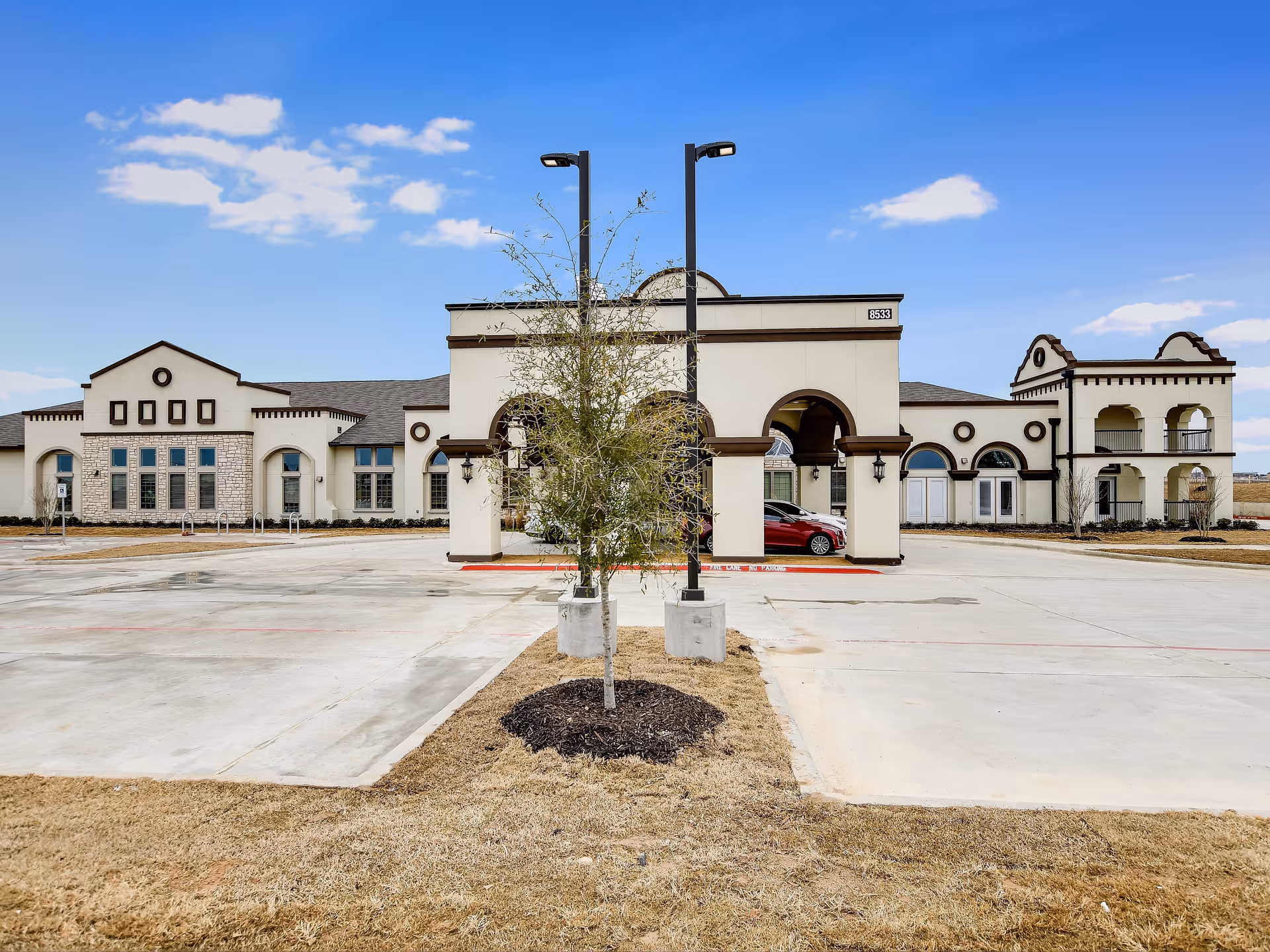 Exterior view of The Grandview of Chisholm Trail building with a clear blue sky, a paved parking area, a small tree in the center foreground, and a red car parked under an archway entrance.