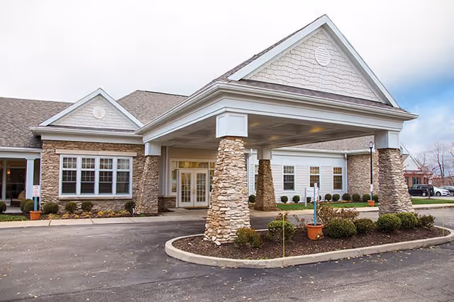 Front exterior view of Richmond Heights Place building with stone pillars supporting a covered entrance, multiple windows, and a landscaped area with shrubs and small plants in front.