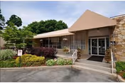 Exterior view of the entrance to Ross Center for Nursing and Rehabilitation, featuring a covered walkway, glass doors, stone wall accents, and surrounding greenery including bushes and trees.