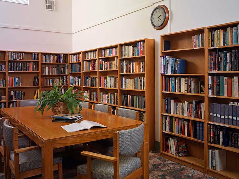 A quiet library room with wooden bookshelves filled with books lining the walls. A wooden table with six cushioned chairs surrounds it, with a potted fern and some open books on the table. A clock is mounted on the wall above the shelves.