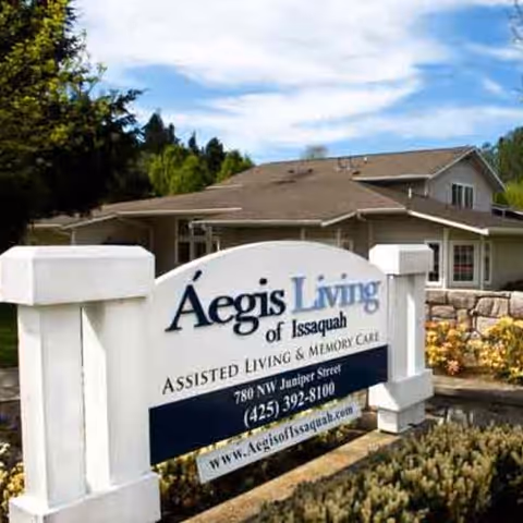 Outdoor view of the Aegis Living of Issaquah facility sign with a building and trees in the background under a partly cloudy sky.