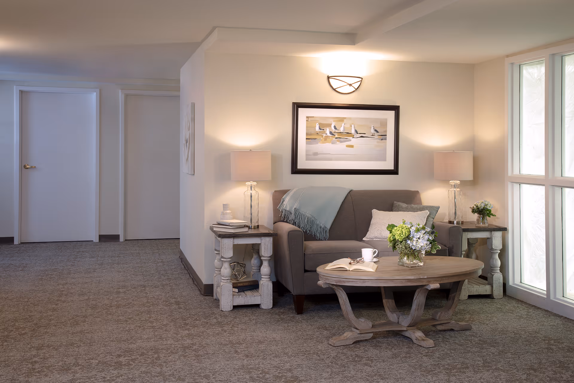 A cozy sitting area in an assisted living facility featuring a gray loveseat with a blue throw blanket and pillows, flanked by two side tables each with a lamp and decorative items. A wooden coffee table in front holds a vase with flowers, an open book, and a cup. The wall behind the loveseat has a framed picture of seagulls on a beach, and there is a large window to the right letting in natural light. The carpeted floor and neutral wall colors create a warm and inviting atmosphere.