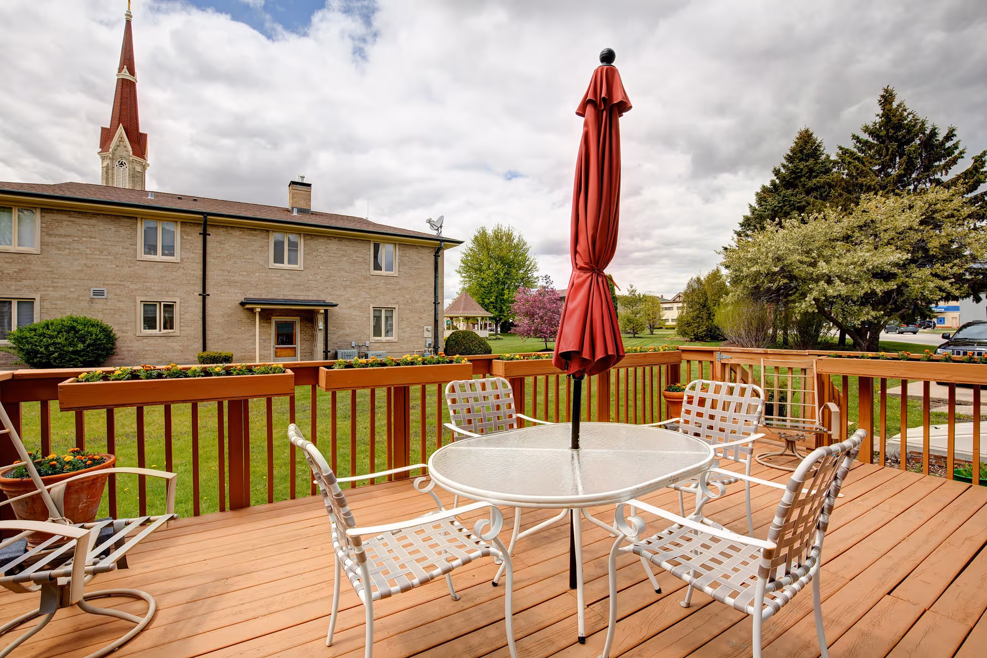 Outdoor wooden deck with a glass-top table and four white metal chairs with woven seats and backs. A closed red patio umbrella is centered on the table. The deck is surrounded by a wooden railing with flower boxes containing orange flowers. In the background, there is a two-story brick building, green grass, trees, and a church steeple under a cloudy sky.