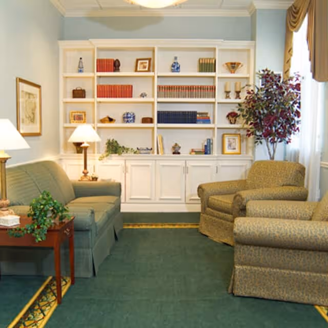 A cozy living room with a green sofa, two patterned armchairs, a wooden side table with a plant, two table lamps, a large white built-in bookshelf filled with books and decorative items, and a tall potted plant near a window with curtains.