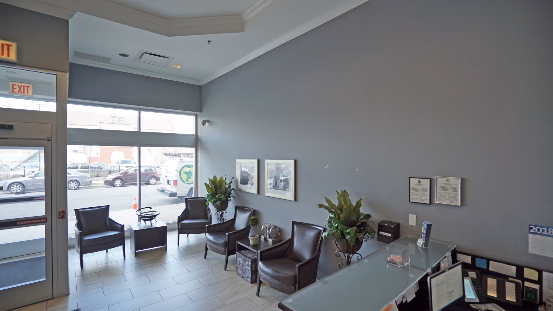 A modern waiting area with four dark leather chairs arranged around two small tables, two large potted plants, framed pictures on a gray wall, and a glass reception desk. Large windows and a glass door provide a view of parked cars outside.