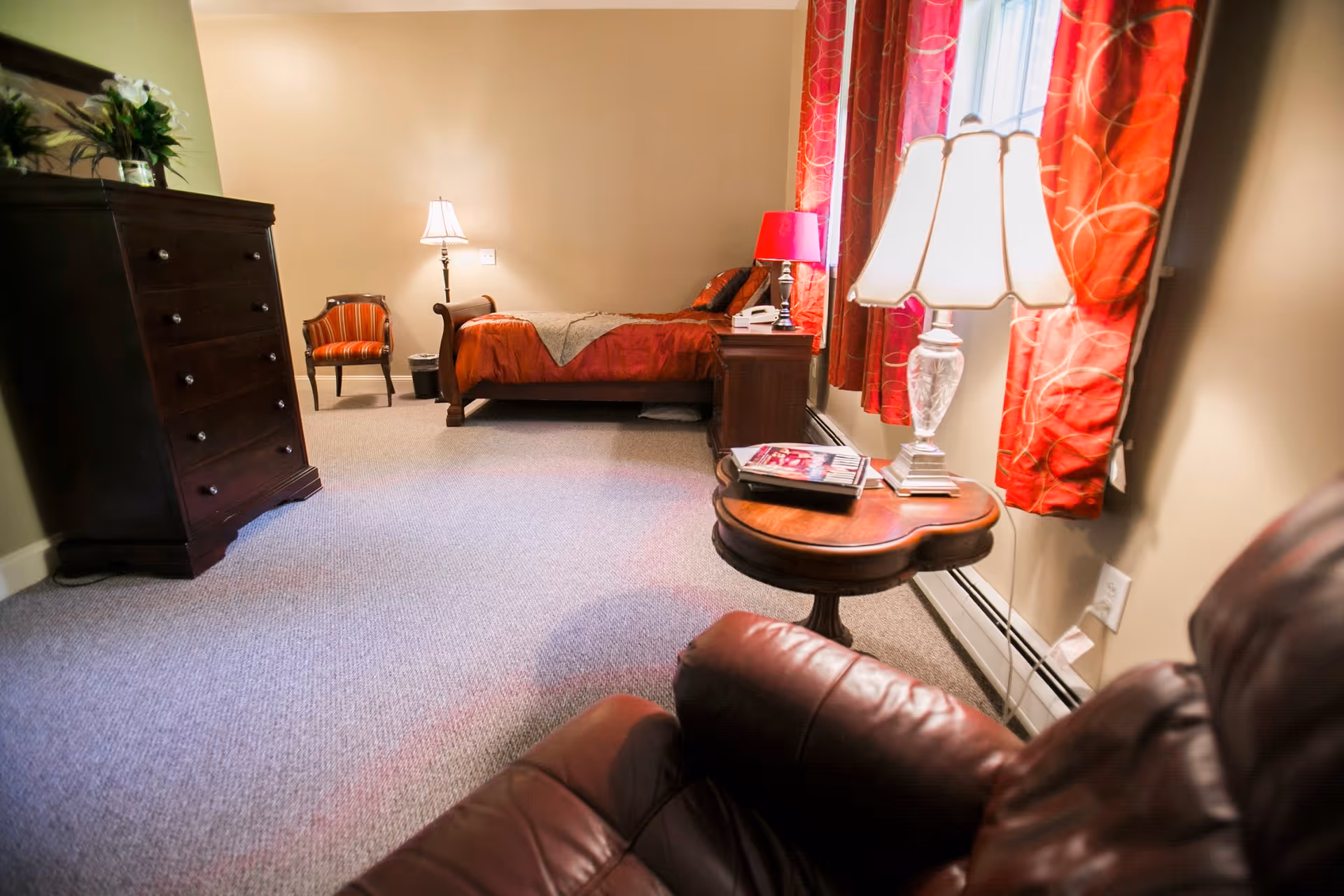 A cozy bedroom in an assisted living facility featuring a bed with red bedding, a wooden dresser, a small round wooden table with a lamp and magazines, a comfortable leather chair, and red curtains on the window.