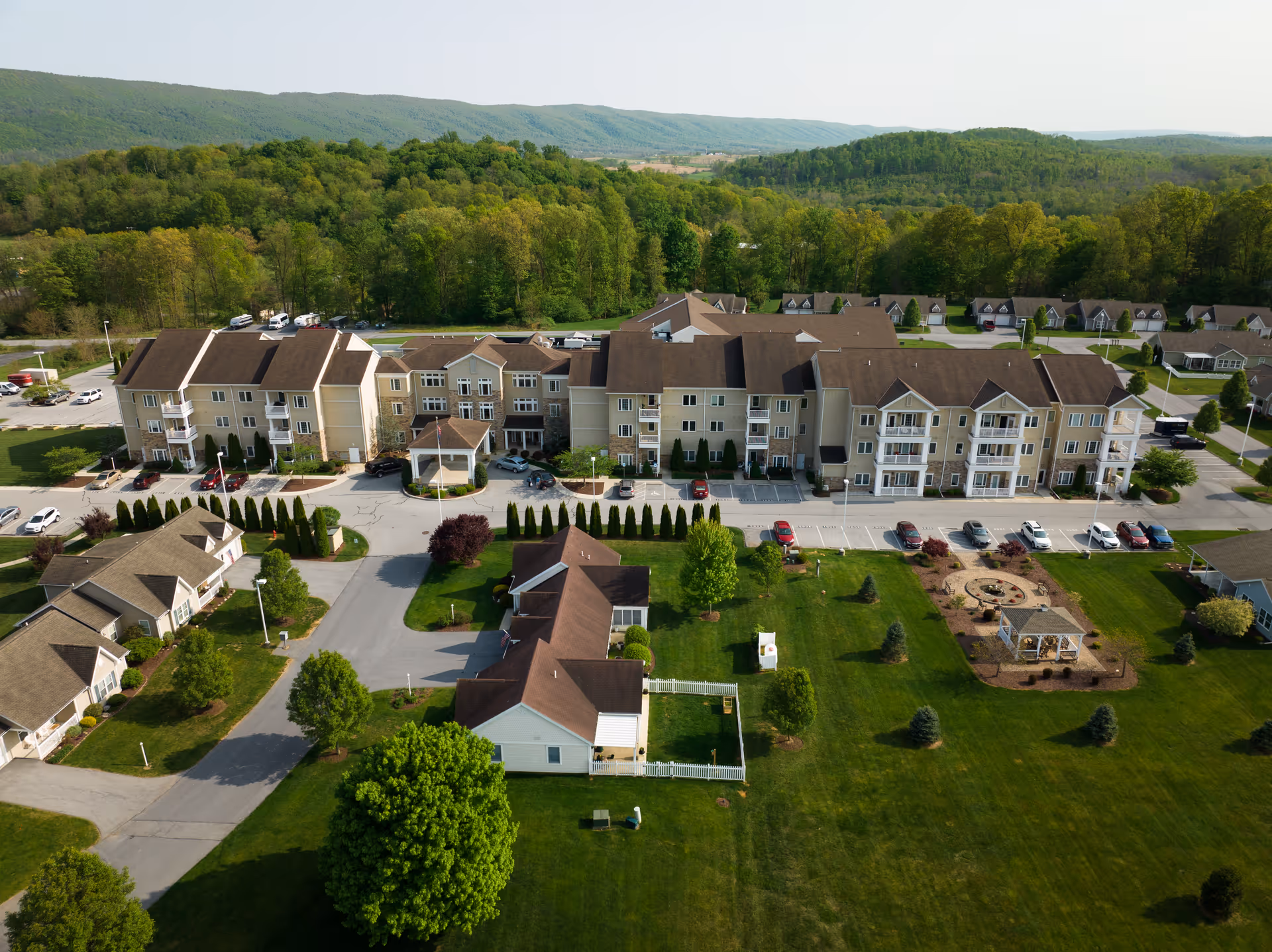 Aerial view of Homewood Living Spring House facility showing a large multi-story residential building with balconies, surrounded by smaller houses, green lawns, trees, and a landscaped garden area with a gazebo. The background features forested hills under a clear sky.