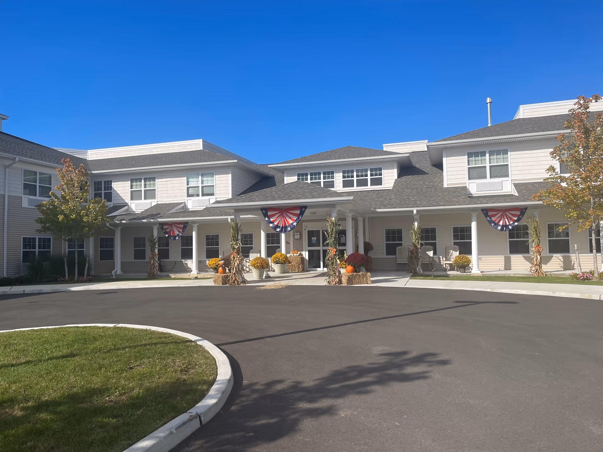 Front exterior view of a two-story senior living facility building with white and beige siding, decorated with patriotic bunting and fall-themed decorations including pumpkins and hay bales. There are trees with autumn foliage on either side of the entrance and a clear blue sky overhead.