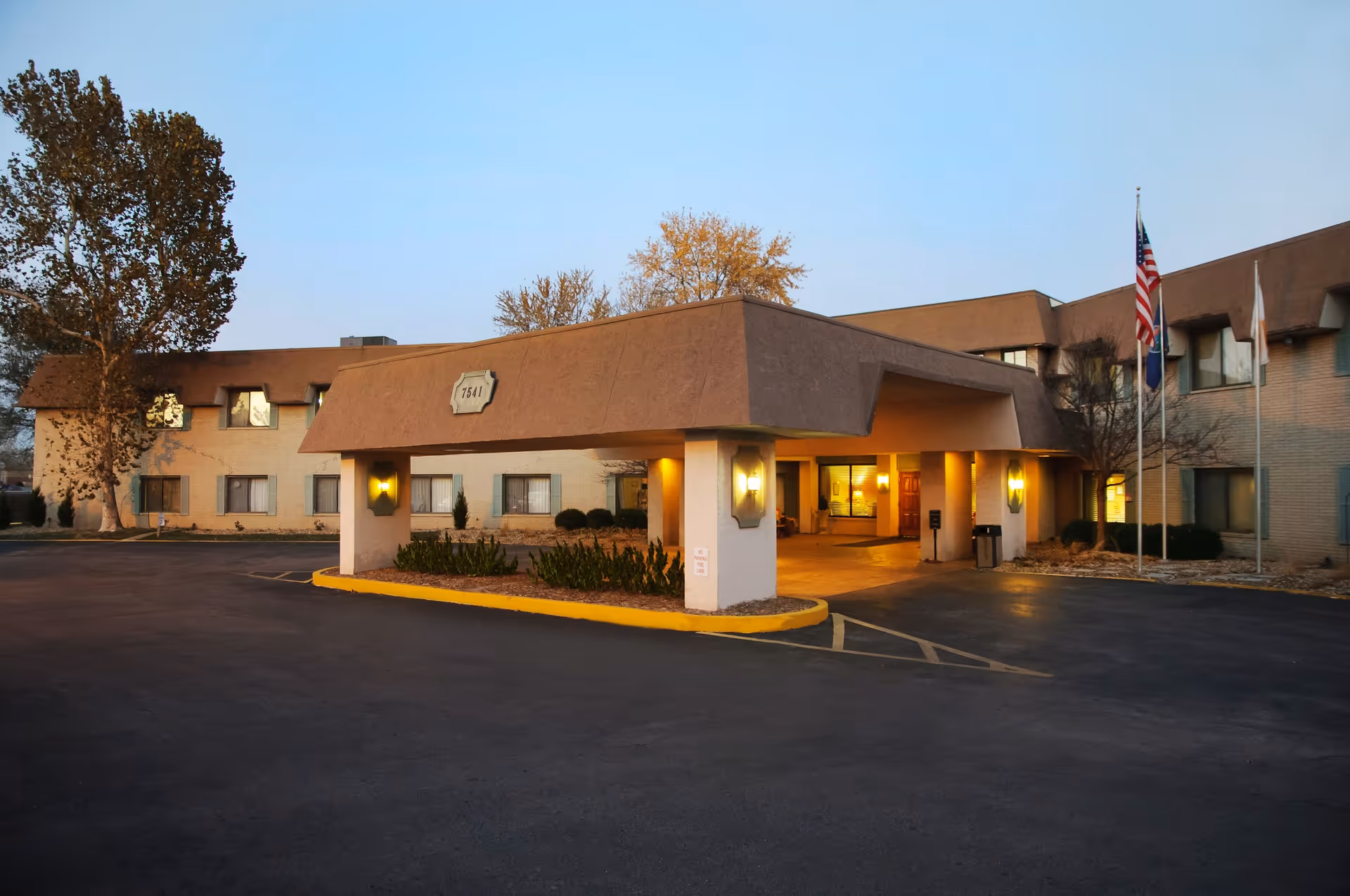 Exterior view of a senior living facility building at dusk with a covered entrance, illuminated wall lights, and three flagpoles displaying the American flag and other flags. The building has two stories with multiple windows and is surrounded by a paved driveway and some trees.
