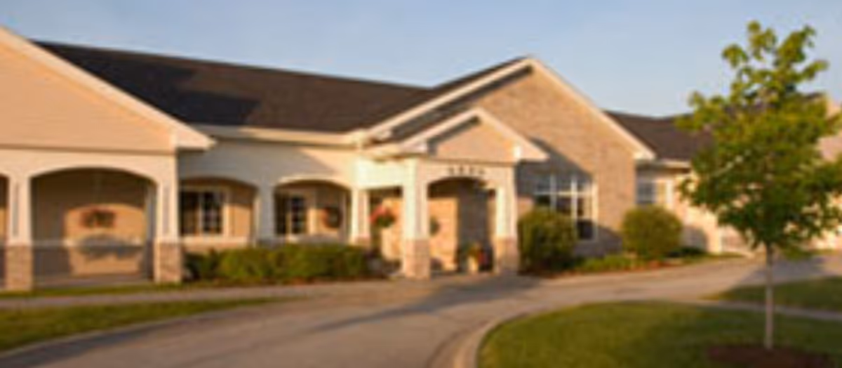 Exterior view of Jersey Ridge Place, a single-story building with a covered entrance, brick and light-colored siding, surrounded by greenery and a curved driveway.