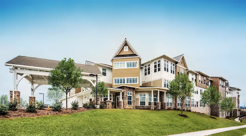 Exterior view of a large, multi-story senior living facility building with beige siding and stone accents, surrounded by landscaped greenery and trees under a clear blue sky.