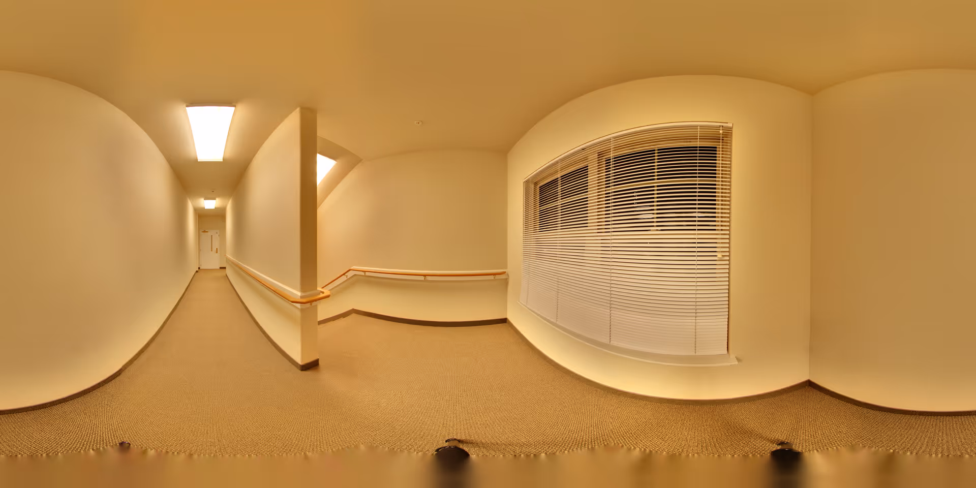 A long, narrow indoor hallway with beige walls and carpeted floor. The hallway features handrails on both sides and a large window with closed blinds on the right. The ceiling has rectangular fluorescent lights, and a door is visible at the far end of the hallway.