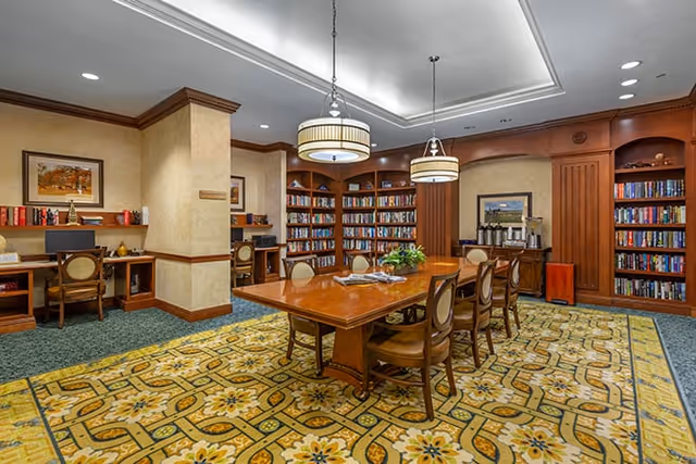 A well-lit library room with wooden bookshelves filled with books along the walls. There is a large wooden table in the center surrounded by chairs. Two pendant lights hang from the ceiling above the table. The room has a patterned carpet and a few framed pictures on the walls. There are also two computer desks with chairs along one wall.