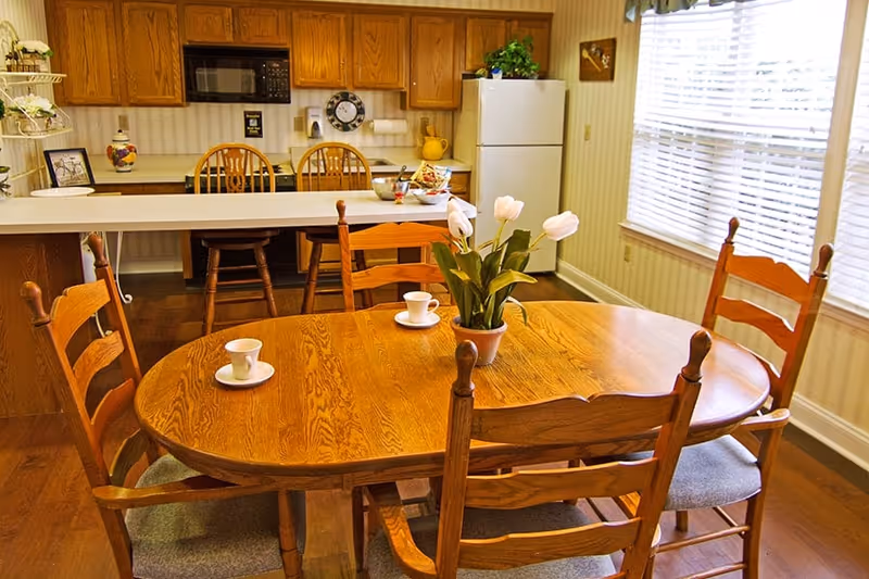 A cozy kitchen and dining area with a wooden oval dining table surrounded by four wooden chairs. On the table are three white cups and saucers and a potted plant with white flowers. Behind the table is a kitchen counter with two wooden bar stools, wooden cabinets, a microwave, a refrigerator, and a window with blinds letting in natural light.