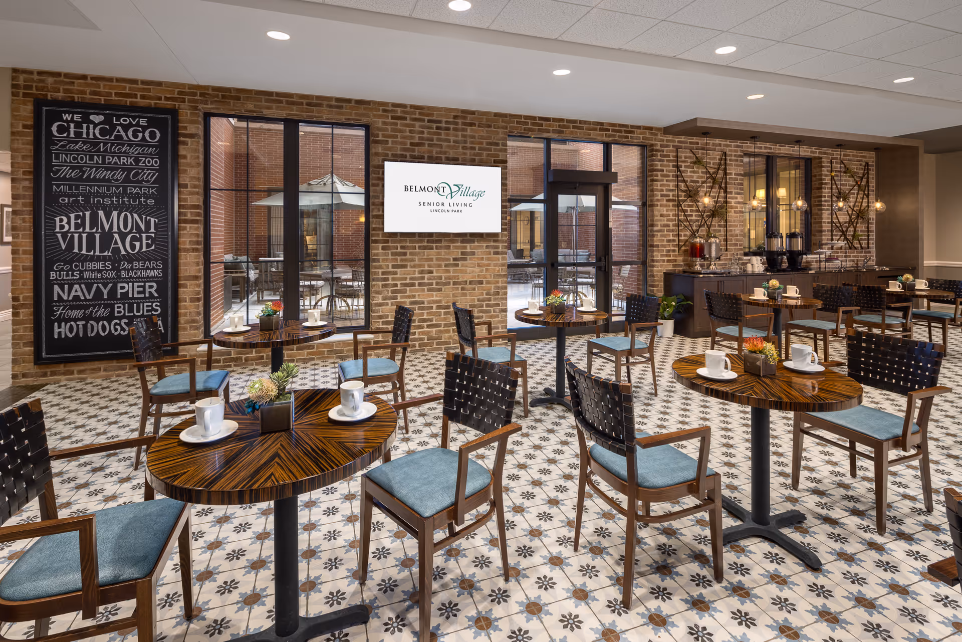 A bright dining area in Belmont Village Senior Living Lincoln Park featuring multiple round wooden tables with blue cushioned chairs. Each table has white coffee cups and small decorative plants. The floor has patterned tiles, and the walls are made of exposed brick. Large windows and glass doors open to an outdoor patio with tables and umbrellas. A sign on the brick wall reads 'Belmont Village Senior Living Lincoln Park,' and a large blackboard-style sign lists Chicago landmarks and sports teams.