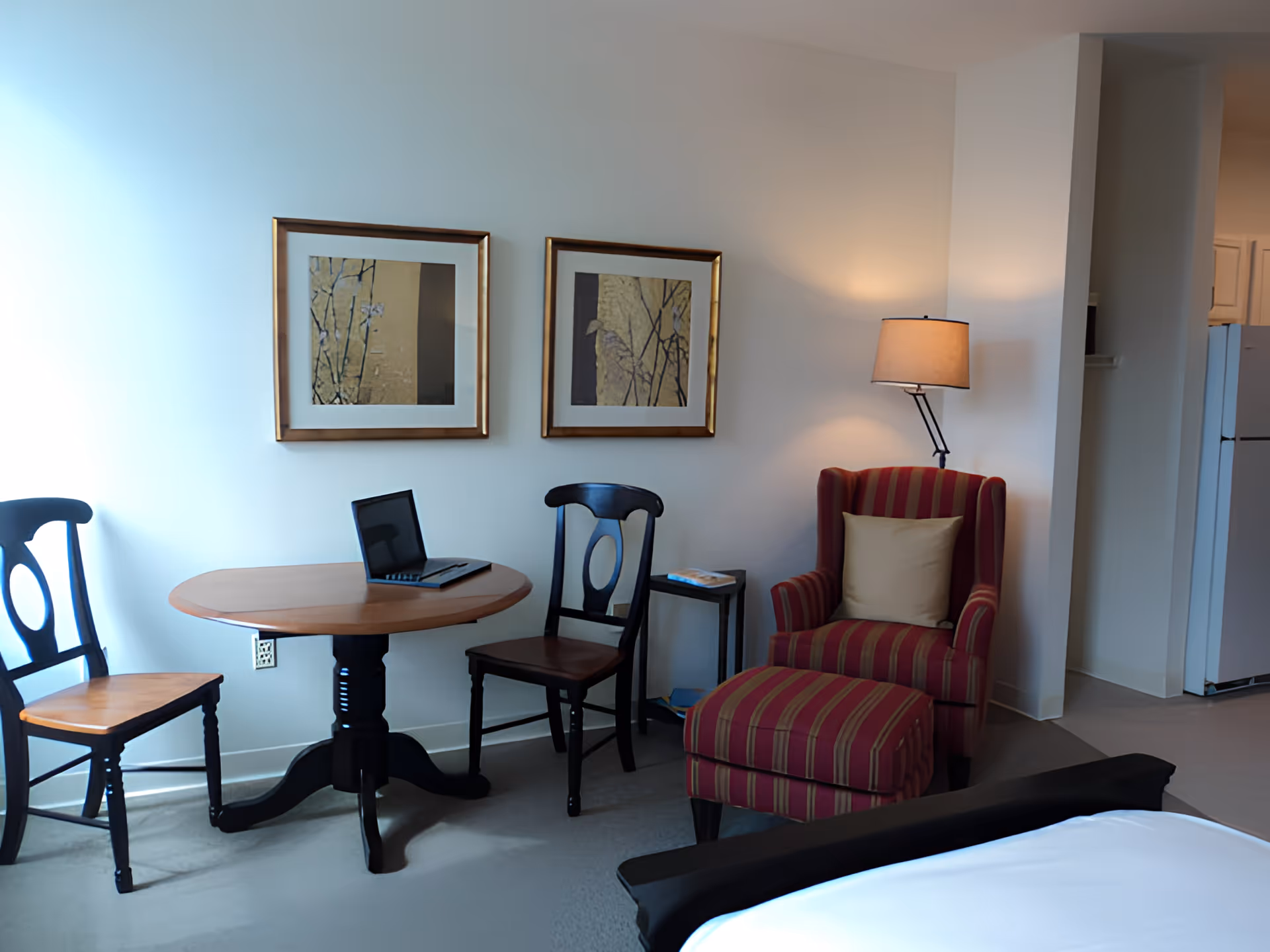 A cozy interior room featuring a small round wooden table with two wooden chairs, a red and beige striped armchair with a matching ottoman, a floor lamp, two framed abstract artworks on the wall, and a partial view of a bed and a refrigerator in the background.