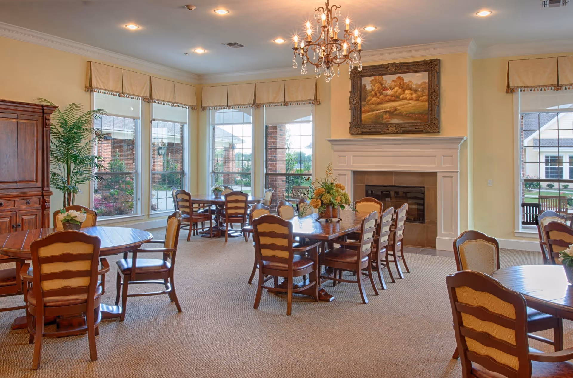 A bright and elegant dining room with multiple wooden tables and chairs arranged neatly. Large windows with beige valances allow natural light to fill the room. A chandelier hangs from the ceiling, and a fireplace with a framed landscape painting above it is visible on one wall. There is a wooden cabinet and a potted plant in the corner.