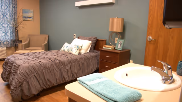A cozy bedroom with a single bed covered in a gray comforter and decorative pillows. Next to the bed is a wooden nightstand with a lamp, books, and a framed photo. A beige armchair is positioned near a window with blue patterned curtains. In the foreground, there is a sink with a chrome faucet and a folded turquoise towel on the counter. The walls are painted a muted teal color.