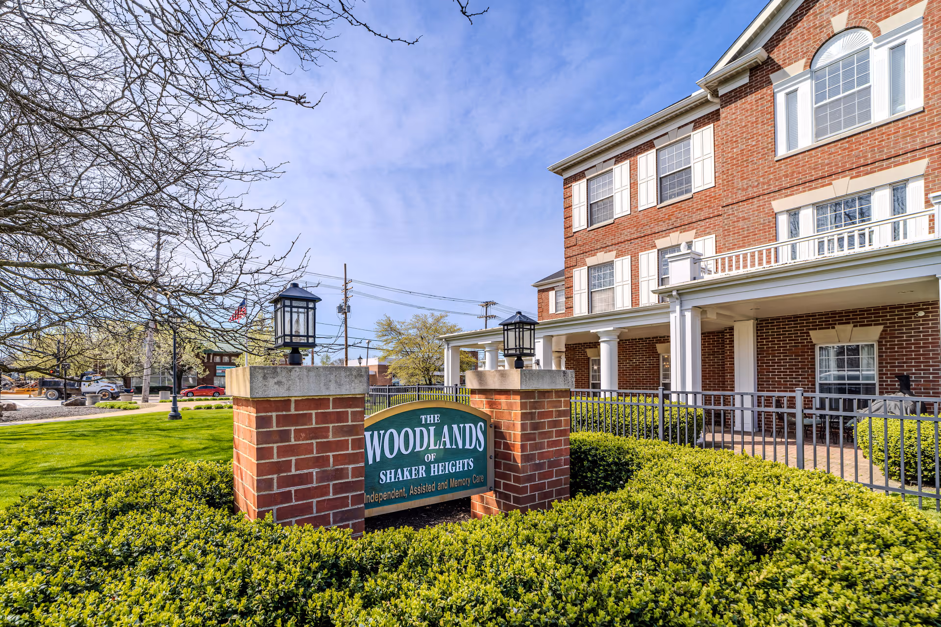 Exterior view of The Woodlands of Shaker Heights senior living facility showing a brick building with white columns and multiple windows. In front of the building is a green sign with white text reading 'The Woodlands of Shaker Heights Independent, Assisted and Memory Care,' surrounded by neatly trimmed bushes and a well-maintained lawn under a partly cloudy sky.